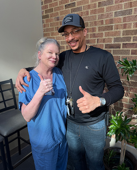 man with black shirt on pictured with hat on it and giving the thumbs up sign with a woman in blue scrubs with a plant in the background